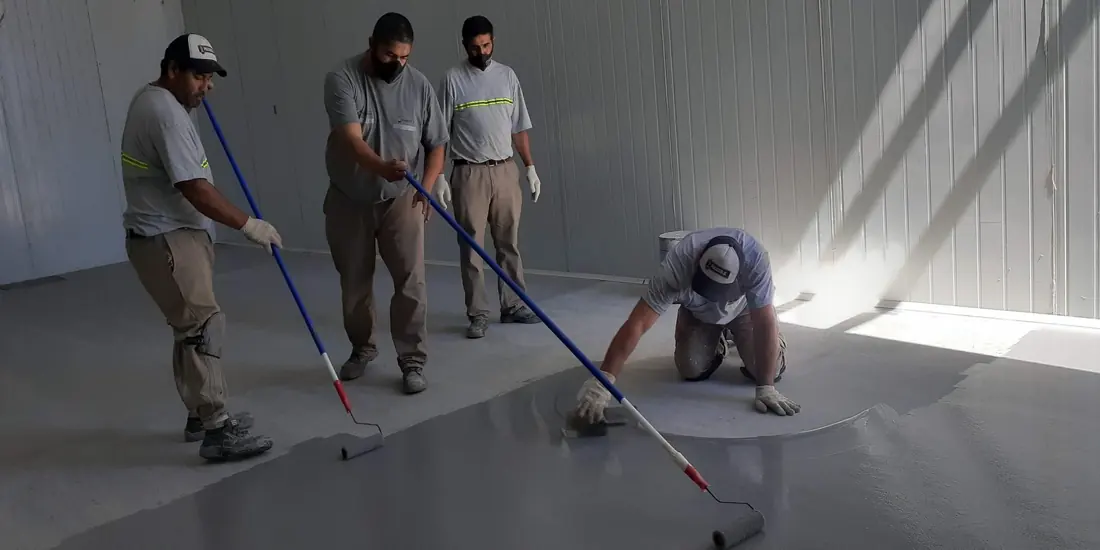 Cuadrilla de cuatro operarios aplicando top coat de poliuretano-cemento con rastrillos en la nave de un frigorífico